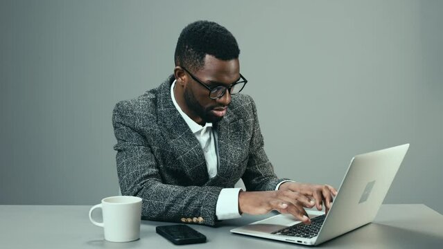 African American businessman working in the office at a laptop with a mug of coffee at his desk on a gray background and tired