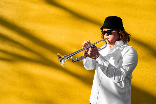 Young Caucasian Long-haired Man In White Shirt, Hat And Sunglasses Playing Funky Jazz On Silver Trumpet With Pleasure Standing Near Yellow Wall On City Street On Concert, Event Or School Exam Outside