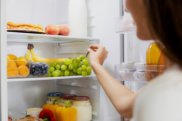 Woman hand taking, grabbing or picks up green bunch of grapes out of open refrigerator shelf or...