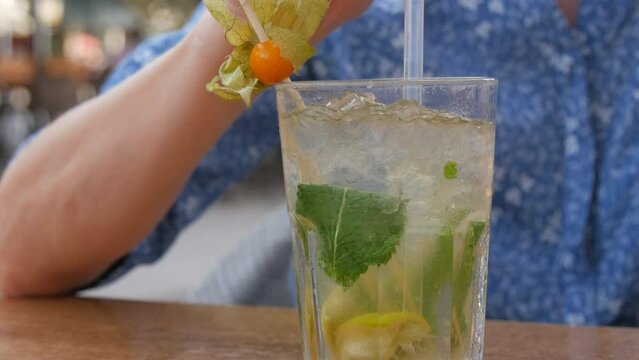 Summer cocktail. Mojito cocktail in a transparent glass glass with pieces of ice, a mint and lemon leaf, a woman drinks a cooling cocktail from a cocktail tube on the veranda of a summer cafe