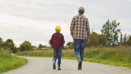 Fototapeta premium Farmer and his son in front of a sunset agricultural landscape. Man and a boy in a countryside field. Fatherhood, country life, farming and country lifestyle.
