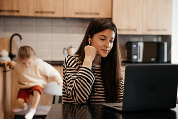 Freelancer woman sits by the table in the home kitchen office, working on laptop. Playful child...