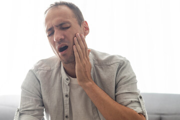 Male patient with severe toothache touching cheek with grimace of pain. Young man with acute dental abscess or inflamed tooth nerve suffering at home. Concept of people having teeth problems.