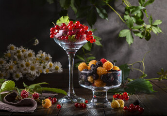 Red currants in a glass wine glass, raspberries and a bouquet of daisies on a wooden table