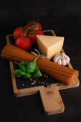 On the board spaghetti, basil, seasonings, garlic, cheese and tomatoes on a dark table. The concept of cooking.