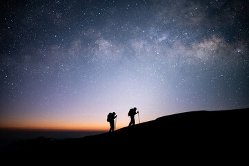 Naklejka na ściany i meble Silhouette of two young traveler and backpacker hiking to the top of the mountain with beautiful view star, milky way over the sky. He enjoyed traveling and was successful when he reached the summit.
