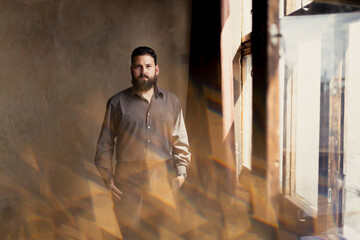 A young man with a beard in a suit is standing by the window