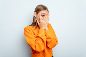 Young caucasian girl isolated on blue background blink through fingers frightened and nervous.