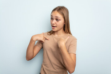 Young caucasian girl isolated on blue background surprised pointing with finger, smiling broadly.