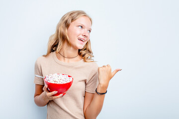 Caucasian teen girl eating cereals isolated on blue background points with thumb finger away, laughing and carefree.