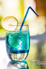 A glass of delicious blue cocktail blue lagoon and yellow lemons on the background of nature. Alcoholic cocktail juicy fruit blue with curacao liqueur, ice cubes and lemon slices.