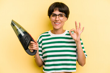 Young caucasian woman holding a hand vacuum cleaner isolated on yellow background cheerful and confident showing ok gesture. © Asier