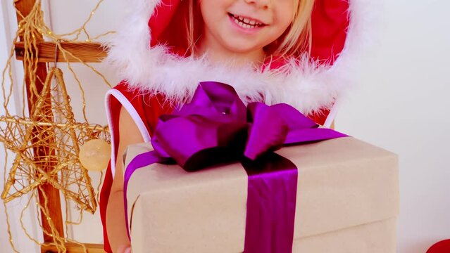 gift with purple ribbon ​​in hands of toddler, small child, blonde girl 3 years old in red New Year's dress close-up, gift from Santa Claus for Christmas, happy childhood, dream of fairy tale, magic