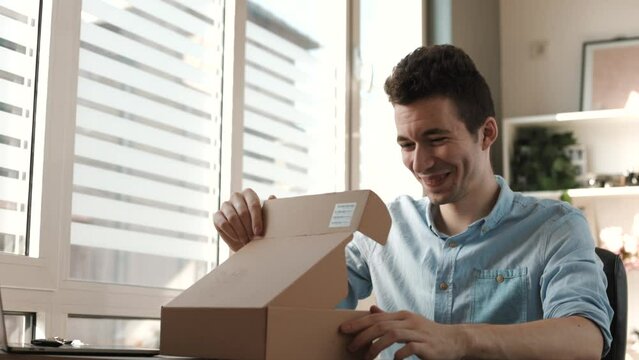 Young smiling man customer opening parcel cardboard box sitting at home office desk. Happy customer

