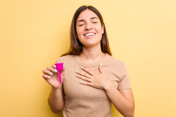 Young caucasian woman holding a menstrual cup isolated on yellow background laughs out loudly keeping hand on chest.