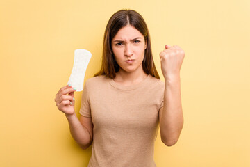 Young caucasian woman holding a sanitary napkin isolated on yellow background showing fist to camera, aggressive facial expression.