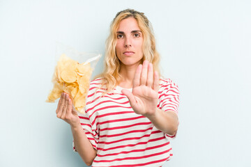 Young caucasian woman holding a bag of chips isolated on blue background standing with outstretched hand showing stop sign, preventing you.
