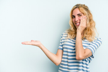 Young caucasian woman isolated on blue background holds copy space on a palm, keep hand over cheek. Amazed and delighted.