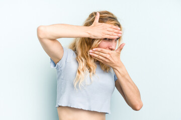 Young caucasian woman isolated on blue background blink at the camera through fingers, embarrassed covering face.