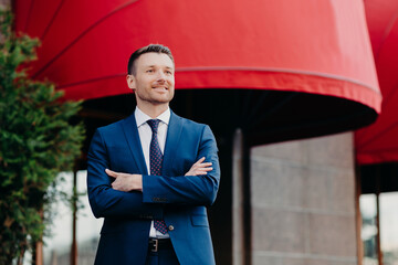 Self assured male CEO in formal suit stands crossed hands, looks with satisfied expression into...