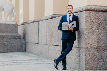 Photo of successful male boss stands crossed legs, holds business magazine, reads necessary information, prepares for presentation in front of colleagues, stands near office building, dressed formally