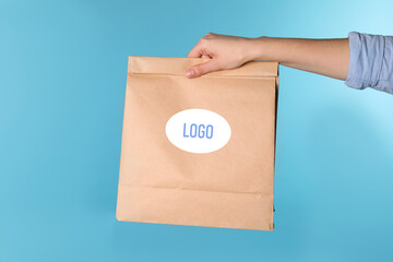 Woman holding paper bag with with logo on light blue background, closeup
