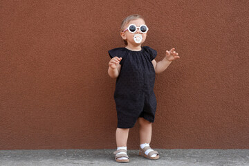 Cute little girl wearing stylish clothes with sunglasses and pacifier near brown wall outdoors
