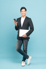 Young asian businessman in smart casual style using smartphone and holding a laptop isolated on blue background