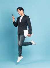 Young asian businessman in smart casual style using smartphone and holding a laptop jumping isolated on blue background