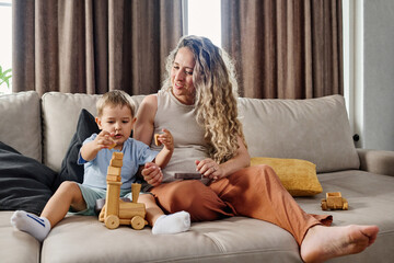 Cute little boy putting stack of wooden cubes on top of toy locomotive while sitting next to his mother on wide comfortable couch