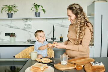 Young careful mother preparing sandwich with honey or jam and passing it to her happy little son sitting in armchair by kitchen table
