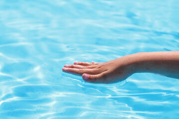 Child's hand touches blue water in the pool. Summer time close-up