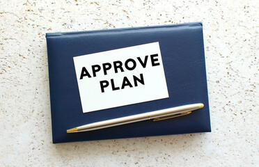 Text APPROVE PLAN on a business card lying on a blue notebook next to the pen.