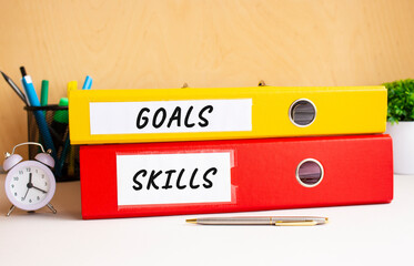 Red and yellow folders lie on the office table next to the clock and pen. Inscriptions on GOALS and SKILLS folders.