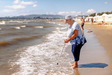 An old woman in a hat with a cane walks to the sea on the beach barefoot