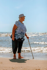 Old woman in a hat with a cane on the beach looks back at the sea while walking barefoot