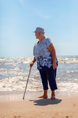 Old woman in a hat with a cane on the beach looks back at the sea while walking barefoot