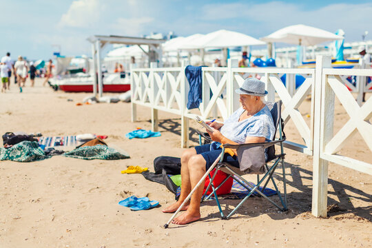 Old woman in a hat sits in a tourist chair on the beach and solves crossword puzzles, a general plan