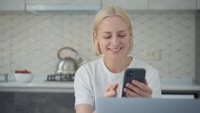 Joyful blonde woman waves bye in video call when sitting at laptop in kitchen