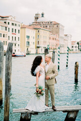 Fototapeta na wymiar Groom hugs bride on a wooden bridge against the backdrop of old houses in Venice