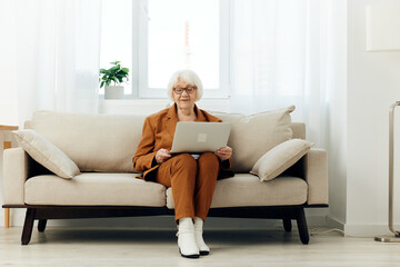 a serious elderly lady is sitting on a beige sofa in a brown suit with a laptop on her lap and enthusiastically working while at home. Full-length photo
