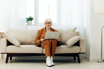 Obraz premium a serious elderly lady is sitting on a beige sofa in a brown suit with a laptop on her lap and enthusiastically working while at home. Full-length photo