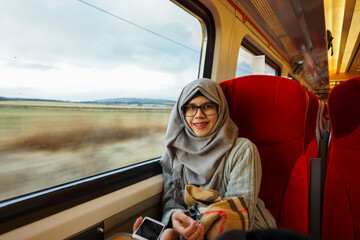 Portrait of young and beautiful Asian Muslim woman wearing eyeglasses and hijab sitting alone against the window in a moving train. Happy and excited expression. 