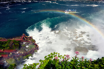 The highest waterfall in the state in summertime colorful strong splash water white form pink flowers foreground over blue sea, yacht boat moving pass beautiful rainbow across the ocean over city