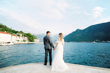 Bride and groom stand holding hands on the pier against the backdrop of Perast. Montenegro