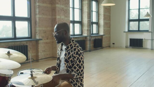 African American man sitting at drum set in spacious loft studio, telling something on camera and then playing drums while giving online music class