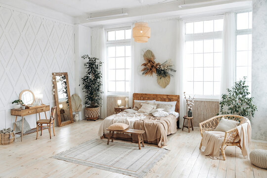 Cozy rustic bedroom with boho ethnic decor. Bright spacious apartment with large windows. Nobody. Wooden furniture. Boudoir table. Large mirror. Handmade textile. Plants in the interior.