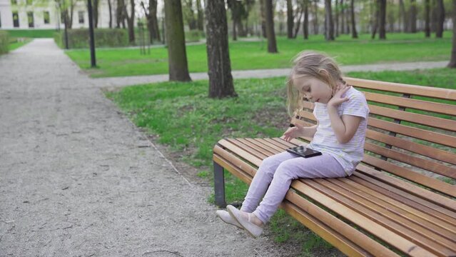 A girl sits on a park bench and ogles a video on her smartphone.