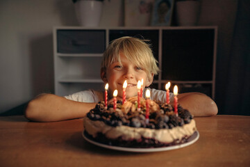 a boy, a child, a teenager, a blonde at the table in front of a cake with lit candles on his...
