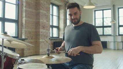 Young bearded man sitting at drum kit, talking on camera and showing drumstick while giving online drum lesson